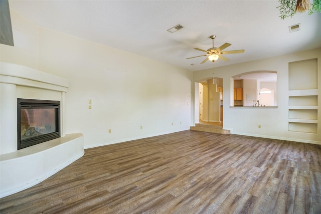 unfurnished living room with visible vents, arched walkways, a glass covered fireplace, ceiling fan, and wood finished floors