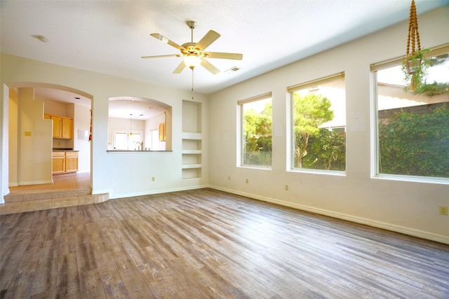 unfurnished living room with a ceiling fan, built in shelves, baseboards, and wood finished floors