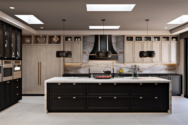 kitchen with tasteful backsplash, glass insert cabinets, dark cabinets, a kitchen island with sink, and ventilation hood