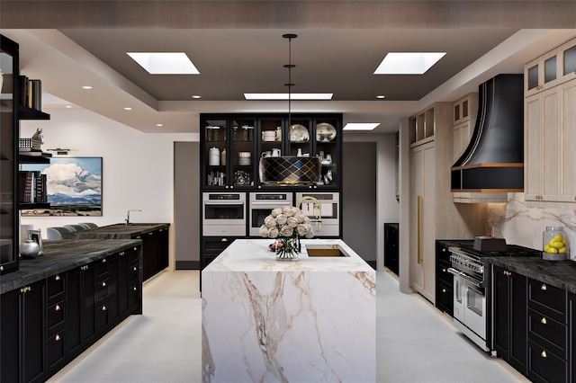 kitchen featuring stainless steel appliances, range hood, dark cabinets, and a skylight