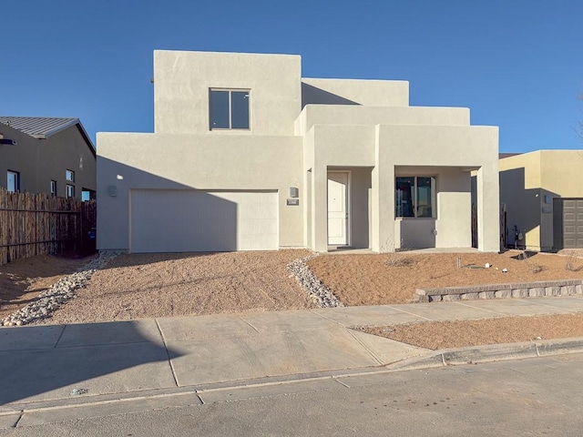 pueblo revival-style home with driveway, an attached garage, fence, and stucco siding