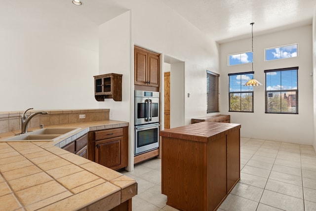 kitchen with light tile patterned floors, tile counters, hanging light fixtures, double oven, and a sink