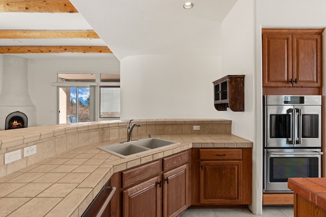 kitchen featuring tile counters, brown cabinets, stainless steel double oven, a fireplace, and a sink