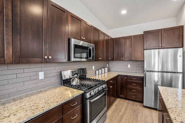 kitchen with appliances with stainless steel finishes, light wood finished floors, decorative backsplash, light stone countertops, and dark brown cabinets