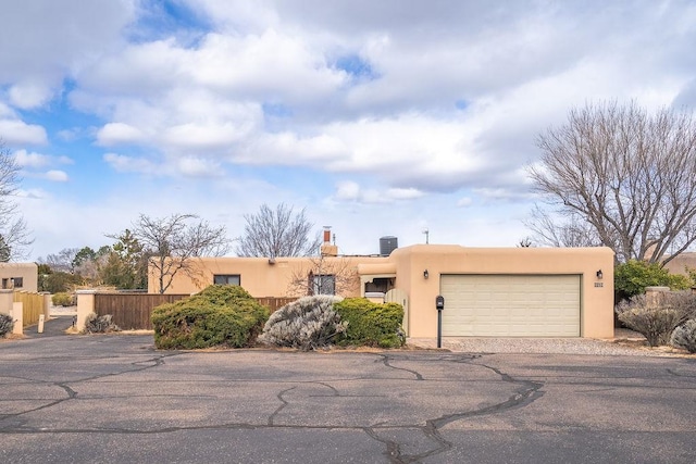 pueblo revival-style home with fence, driveway, an attached garage, a chimney, and stucco siding