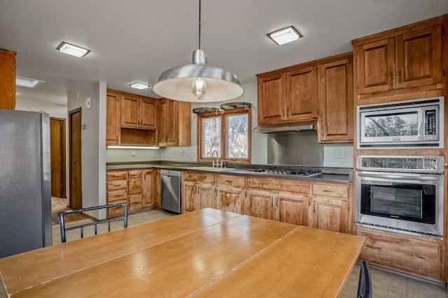 kitchen with a sink, under cabinet range hood, appliances with stainless steel finishes, decorative light fixtures, and brown cabinets