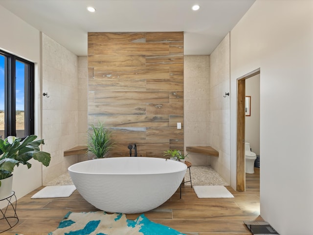 bathroom featuring tile walls, a soaking tub, toilet, and wood finished floors