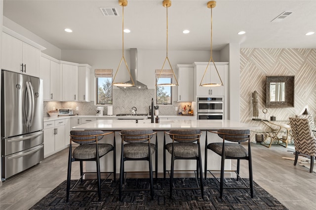 kitchen with visible vents, light countertops, stainless steel appliances, wall chimney exhaust hood, and a kitchen island with sink