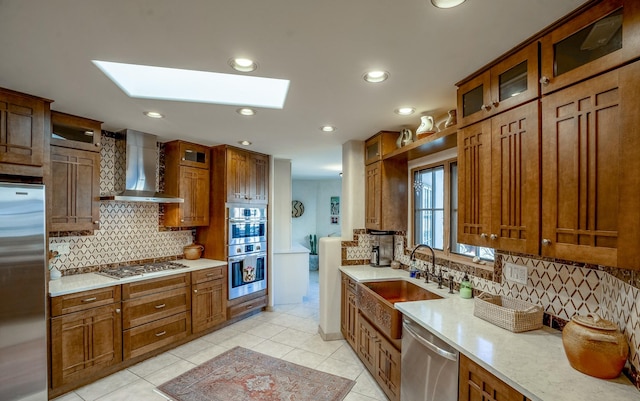 kitchen with wall chimney range hood, light tile patterned floors, appliances with stainless steel finishes, a skylight, and a sink