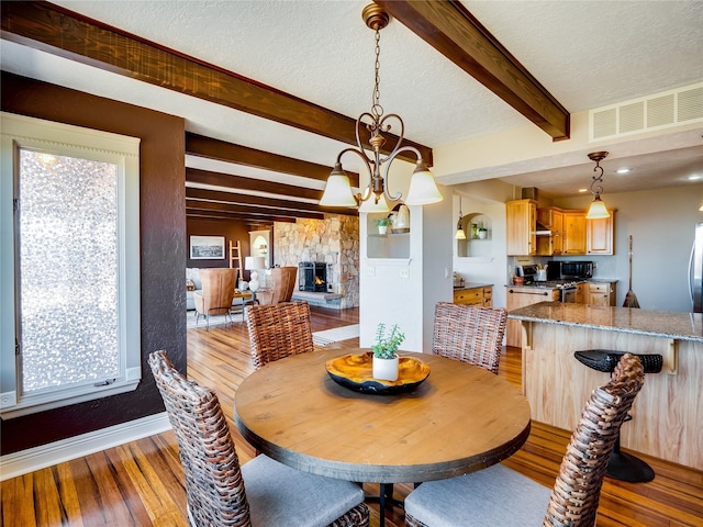 dining area with visible vents, light wood-style flooring, a stone fireplace, a textured ceiling, and beamed ceiling