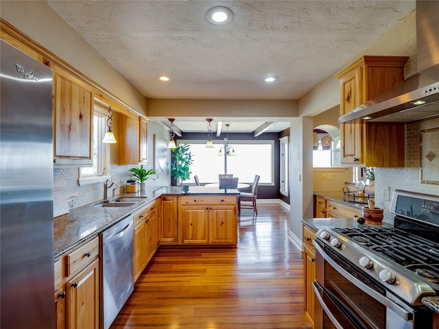 kitchen with wood finished floors, a peninsula, a sink, appliances with stainless steel finishes, and wall chimney range hood