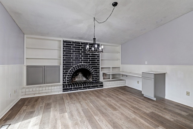 unfurnished living room featuring built in shelves, wood finished floors, wainscoting, a large fireplace, and a chandelier