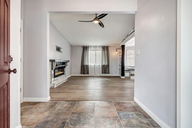 unfurnished living room with a glass covered fireplace, stone finish floor, a ceiling fan, and baseboards