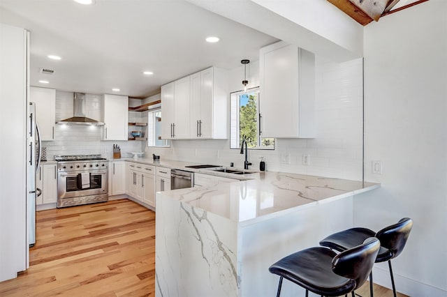 kitchen featuring light stone countertops, a peninsula, stainless steel appliances, wall chimney exhaust hood, and light wood-type flooring