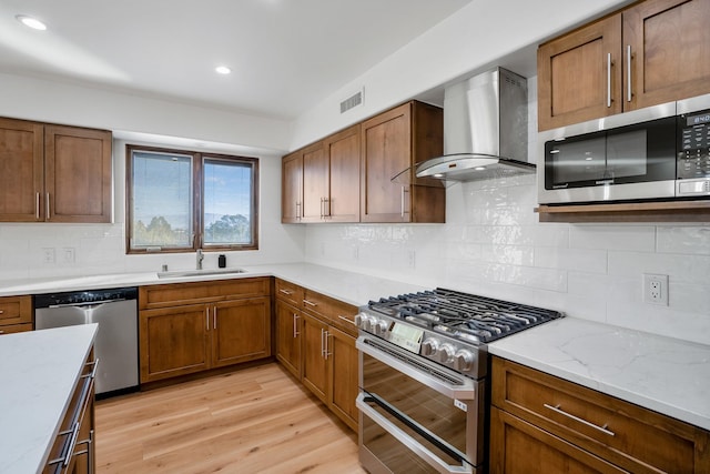 kitchen with wall chimney range hood, appliances with stainless steel finishes, light wood-style floors, brown cabinetry, and a sink