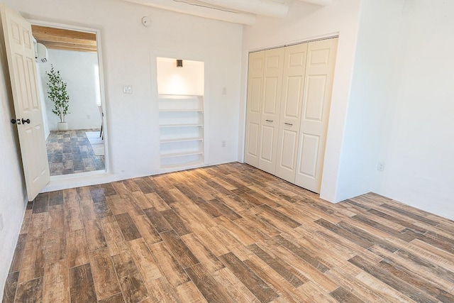 unfurnished bedroom with a closet, beamed ceiling, a wall mounted air conditioner, and wood finished floors