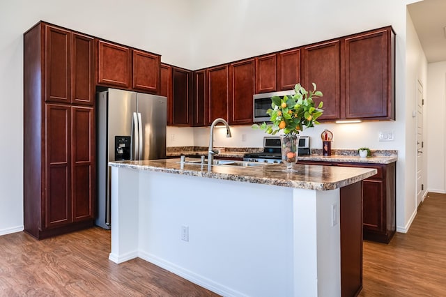 kitchen featuring wood finished floors, a sink, baseboards, stainless steel appliances, and an island with sink