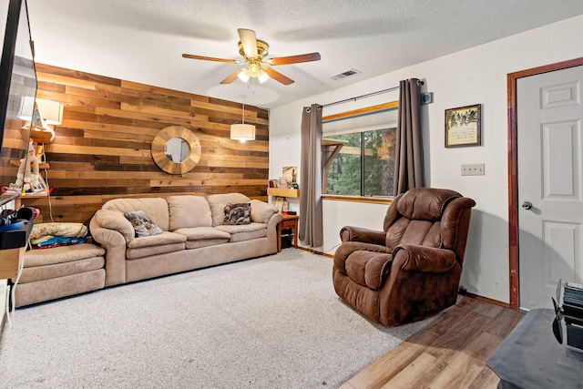 living room with a textured ceiling, wood walls, visible vents, ceiling fan, and wood finished floors