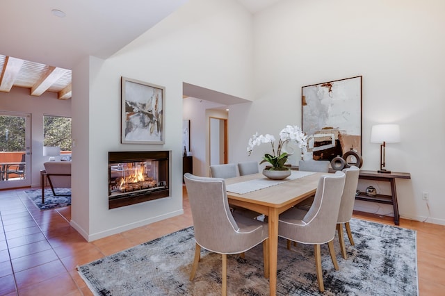 dining room featuring beamed ceiling, light tile patterned flooring, baseboards, and a multi sided fireplace