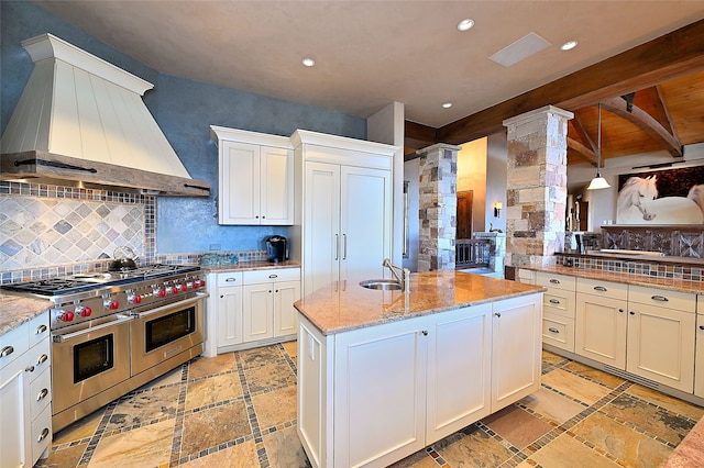 kitchen with range with two ovens, a kitchen island with sink, a sink, decorative columns, and custom exhaust hood