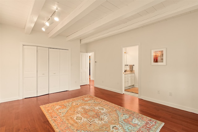 unfurnished bedroom with wood finished floors, a closet, beamed ceiling, baseboards, and ensuite bath