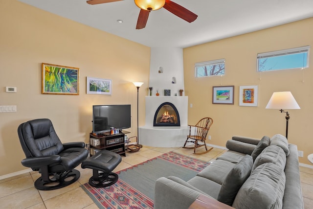 tiled living area with ceiling fan, a warm lit fireplace, and baseboards