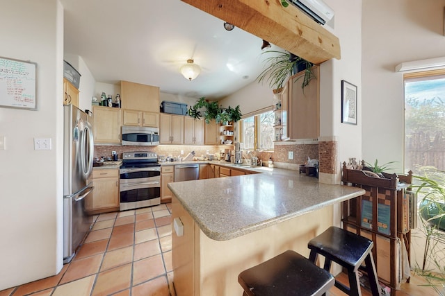kitchen with light brown cabinetry, backsplash, a peninsula, appliances with stainless steel finishes, and a breakfast bar area