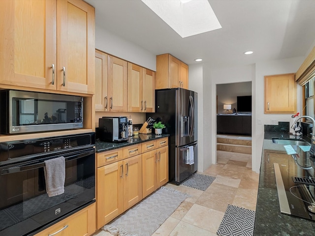 kitchen featuring light brown cabinets, a sink, a skylight, dark stone counters, and black appliances