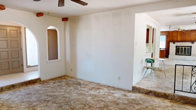 unfurnished living room featuring arched walkways, a brick fireplace, carpet, and ceiling fan
