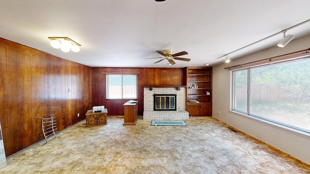 unfurnished living room with wooden walls, ceiling fan, carpet floors, rail lighting, and a brick fireplace
