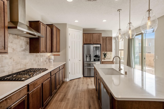 kitchen featuring a sink, light wood-style floors, wall chimney exhaust hood, appliances with stainless steel finishes, and an island with sink