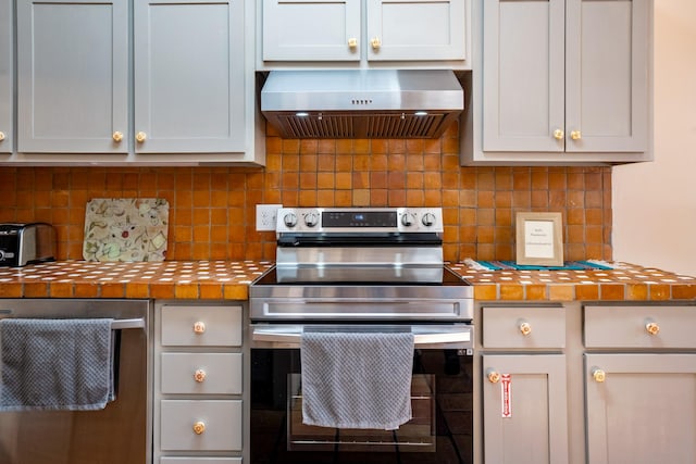 kitchen featuring tile countertops, appliances with stainless steel finishes, wall chimney range hood, and tasteful backsplash