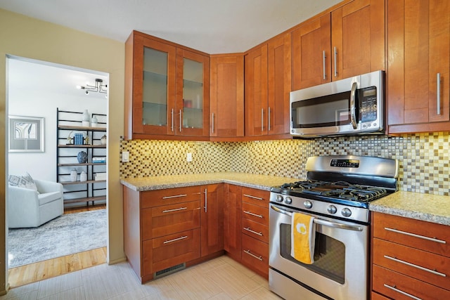 kitchen with brown cabinetry, appliances with stainless steel finishes, tasteful backsplash, and glass insert cabinets