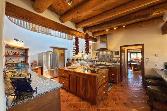 kitchen with wall chimney range hood, beam ceiling, high end refrigerator, tasteful backsplash, and glass insert cabinets