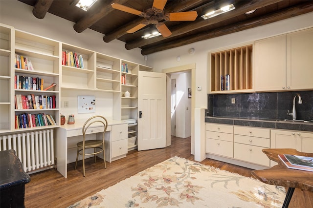 home office with ceiling fan, a sink, radiator heating unit, wood finished floors, and beamed ceiling