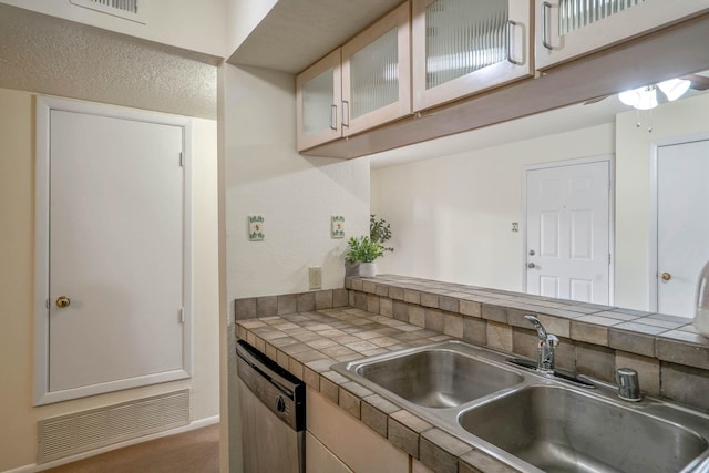 kitchen featuring visible vents, glass insert cabinets, dishwashing machine, tile counters, and a sink