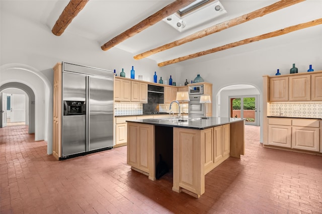 kitchen featuring stainless steel appliances, arched walkways, brick floors, beam ceiling, and tasteful backsplash
