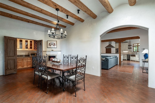 1050 Sierra Del Norte, Santa Fe, NM 87501 - dining room with arched walkways, a warm lit fireplace, beamed ceiling, brick flooring, and a chandelier
