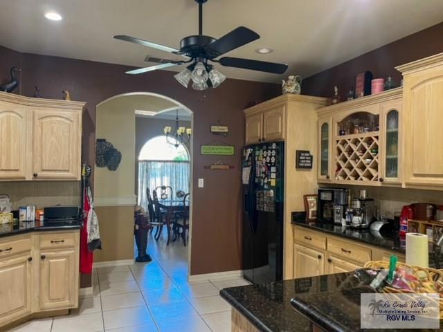 kitchen with light brown cabinets, ceiling fan with notable chandelier, black fridge, dark stone countertops, and light tile patterned floors