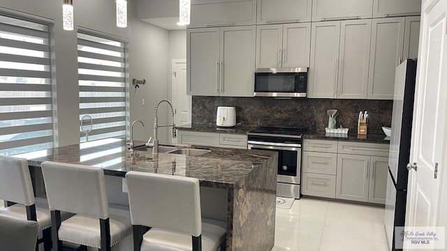 kitchen with gray cabinetry, stainless steel appliances, a sink, hanging light fixtures, and dark stone counters