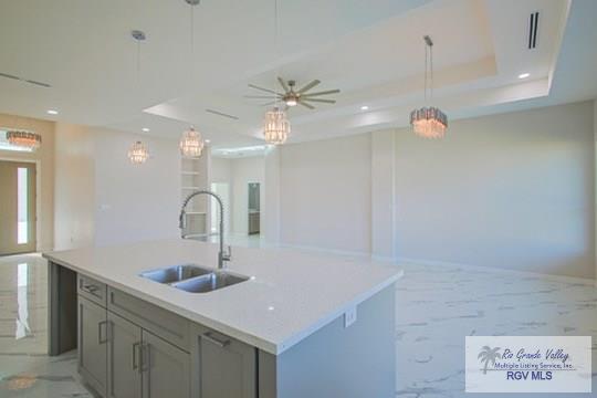 kitchen with gray cabinetry, ceiling fan with notable chandelier, a raised ceiling, sink, and an island with sink