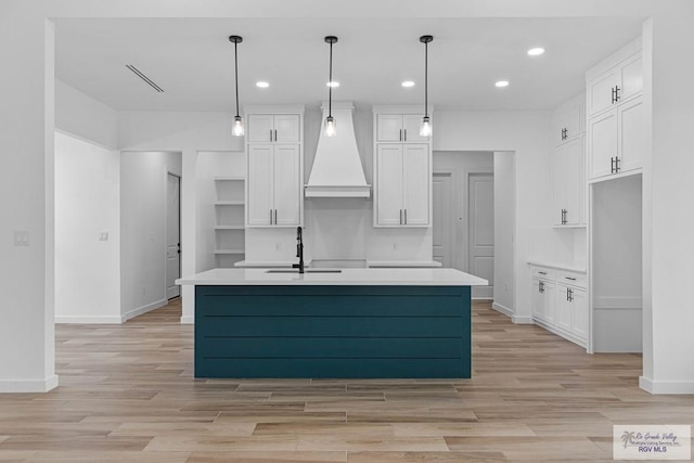 kitchen with pendant lighting, custom exhaust hood, a center island with sink, sink, and white cabinetry