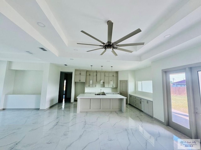 kitchen with pendant lighting, gray cabinetry, a raised ceiling, ceiling fan, and an island with sink