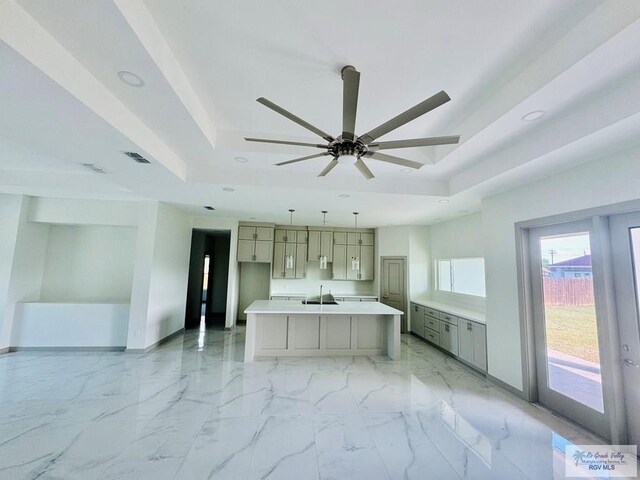 kitchen with pendant lighting, gray cabinetry, a raised ceiling, ceiling fan, and an island with sink