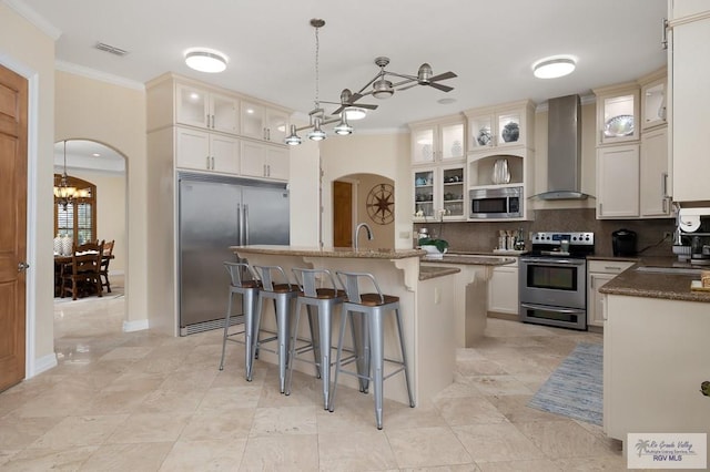 kitchen featuring wall chimney exhaust hood, ceiling fan with notable chandelier, a kitchen island with sink, built in appliances, and hanging light fixtures