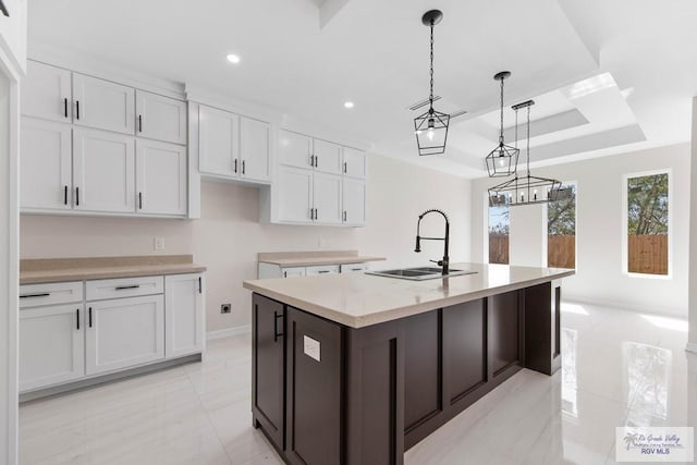 kitchen with a raised ceiling, an island with sink, light countertops, white cabinetry, and a sink