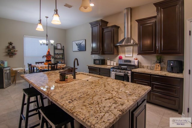 kitchen with stainless steel gas range oven, wall chimney range hood, sink, an island with sink, and dark brown cabinetry