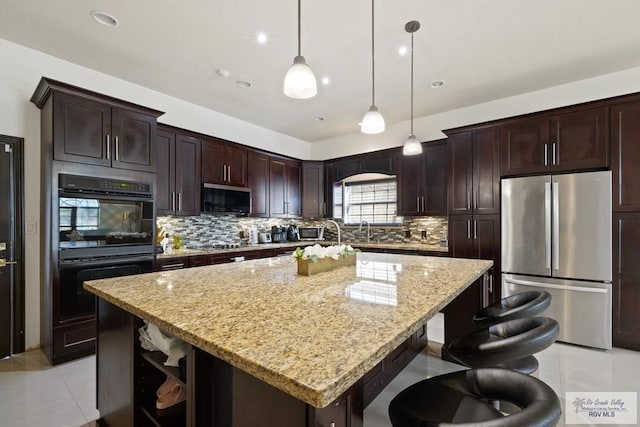 kitchen featuring dark brown cabinetry, stainless steel appliances, light stone counters, backsplash, and an island with sink