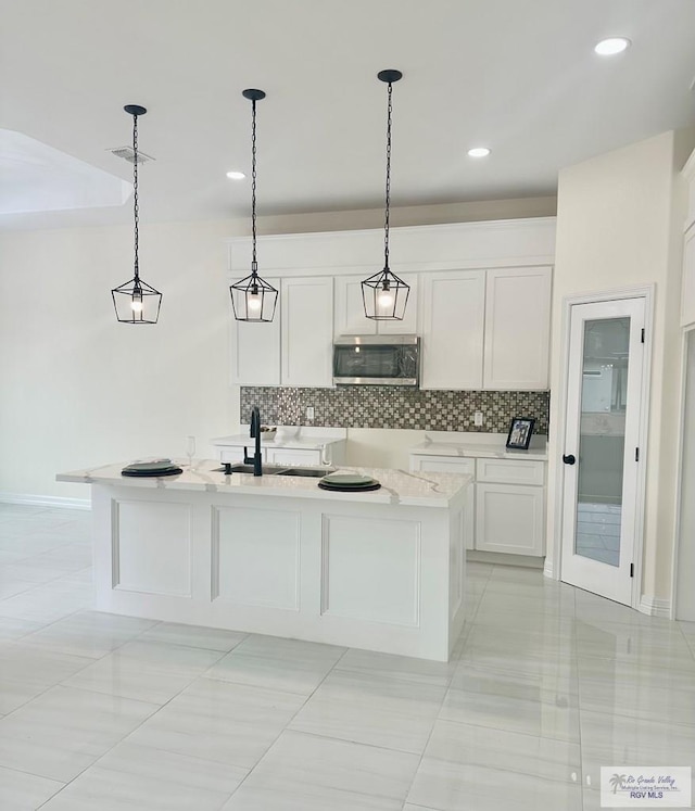 kitchen featuring stainless steel microwave, hanging light fixtures, white cabinetry, a sink, and an island with sink