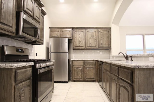 kitchen featuring stainless steel appliances, backsplash, a sink, dark brown cabinets, and light stone countertops